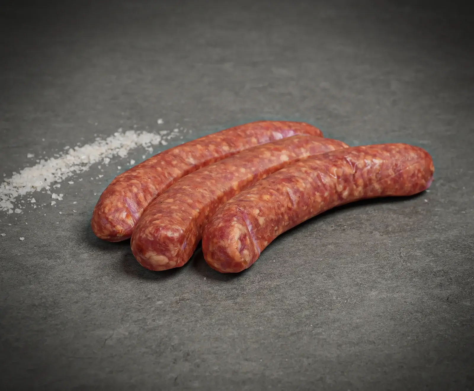 Drei frische, rohe Würste liegen diagonal auf einer dunklen Oberfläche, daneben eine Spur grobes Salz.