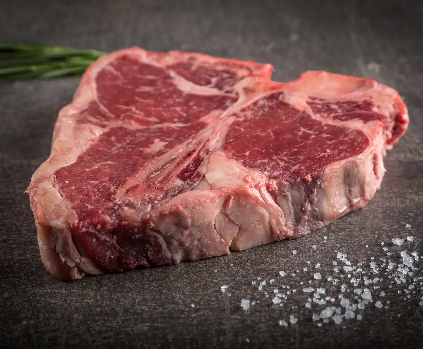 Ein rohes, marmoriertes T-Bone-Steak liegt auf einer dunklen Oberfläche. Im Hintergrund sind Kräuter, im Vordergrund grobes Salz verstreut.