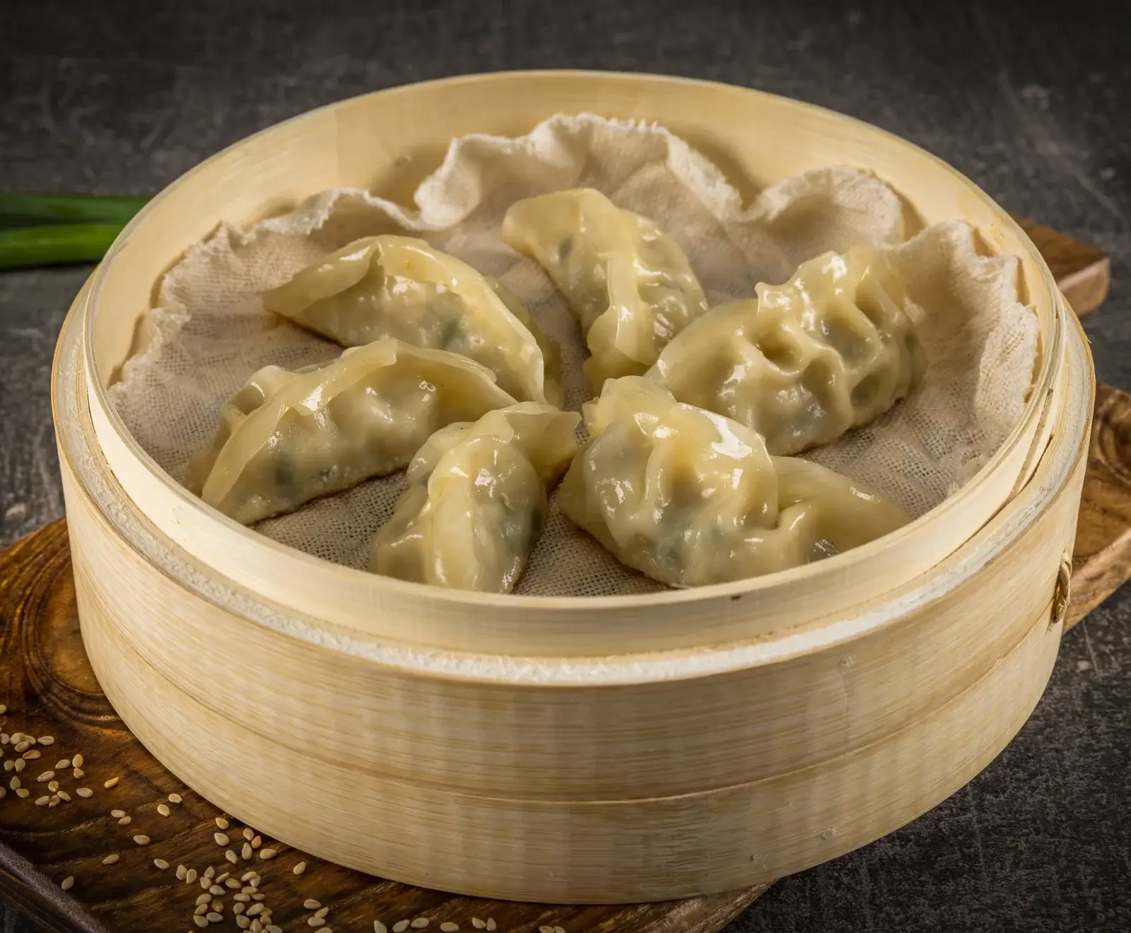 Mehrere glänzende, gedämpfte Teigtaschen in einem Bambusdämpfer auf einem Holzbrett. Im Hintergrund sind Lauchzwiebeln und verstreute Sesamsamen zu sehen.