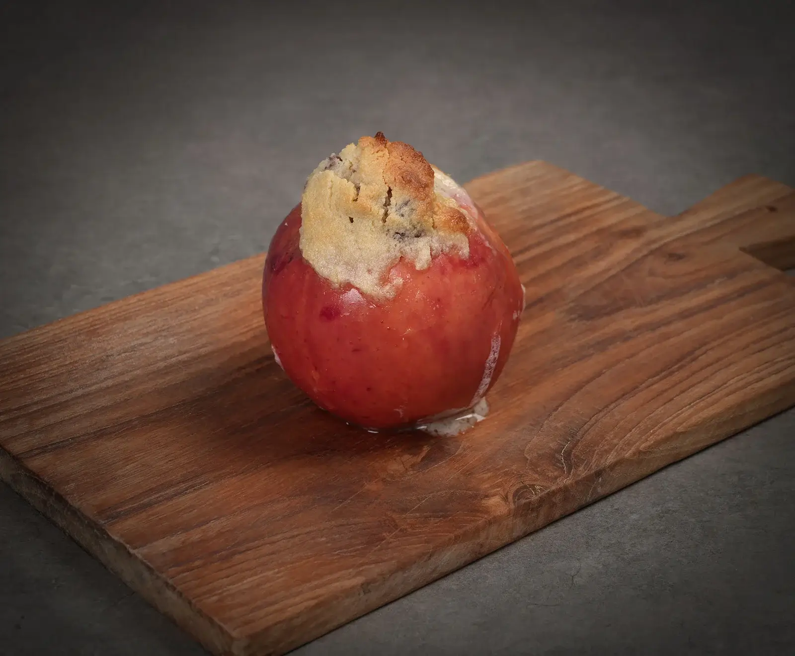 Ein Bratapfel mit goldbrauner Streuselhaube auf einem Holzschneidebrett. Etwas weiße Flüssigkeit ist aus dem Apfel ausgetreten. Dunkelgrauer Hintergrund.
