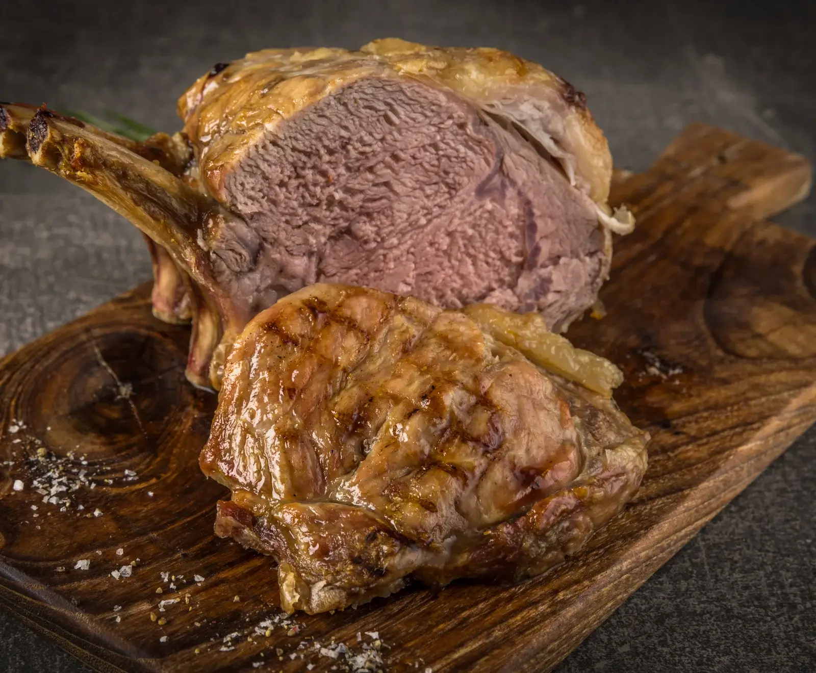 Ein großes, gebratenes Fleischstück mit Knochen, innen rosa gegart, liegt auf einem rustikalen Holzbrett. Davor ein kleineres, gegrilltes Stück. Grobes Salz und Pfeffer sind auf dem Brett verstreut.