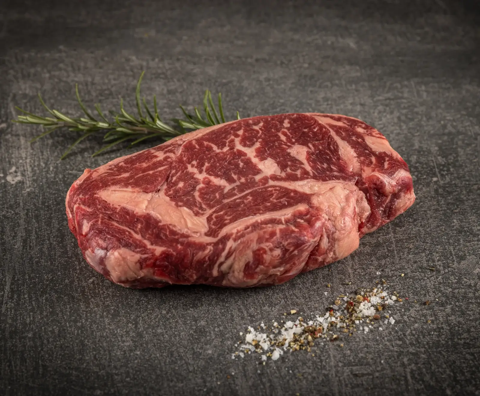 Ein rohes, stark marmoriertes Ribeye-Steak liegt auf einem dunklen Untergrund, daneben ein Rosmarinzweig und eine Prise Salz und Pfeffer.