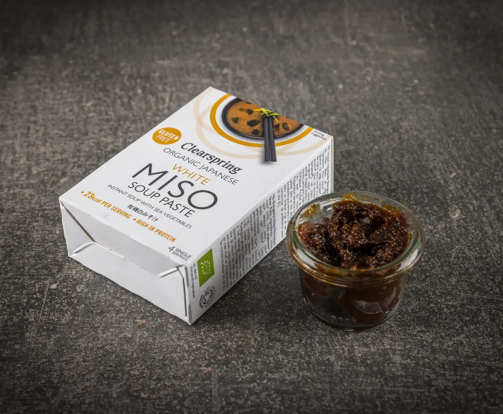 Eine weiße Packung Clearspring Bio Japanische Weiße Miso Suppenpaste und ein kleines Glas mit dunkler Miso-Paste auf einem dunklen, strukturierten Hintergrund.