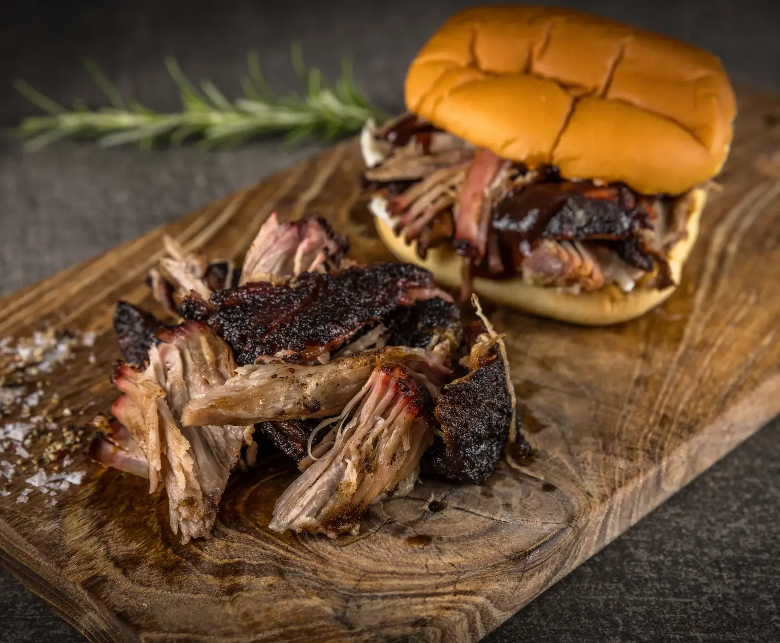 Geschnittenes Schweinefleisch mit dunkler Kruste, serviert auf einem Holzbrett mit Rosmarin und Brötchen im Hintergrund.