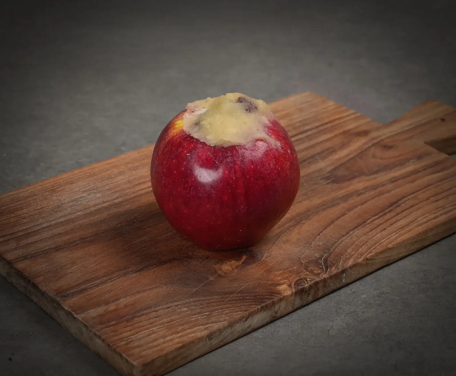 Ein roter Apfel mit ausgehöhltem Kern, gefüllt mit einer hellen Masse und dunklen Stückchen, liegt auf einem rustikalen Holzschneidebrett vor dunklem Hintergrund.
