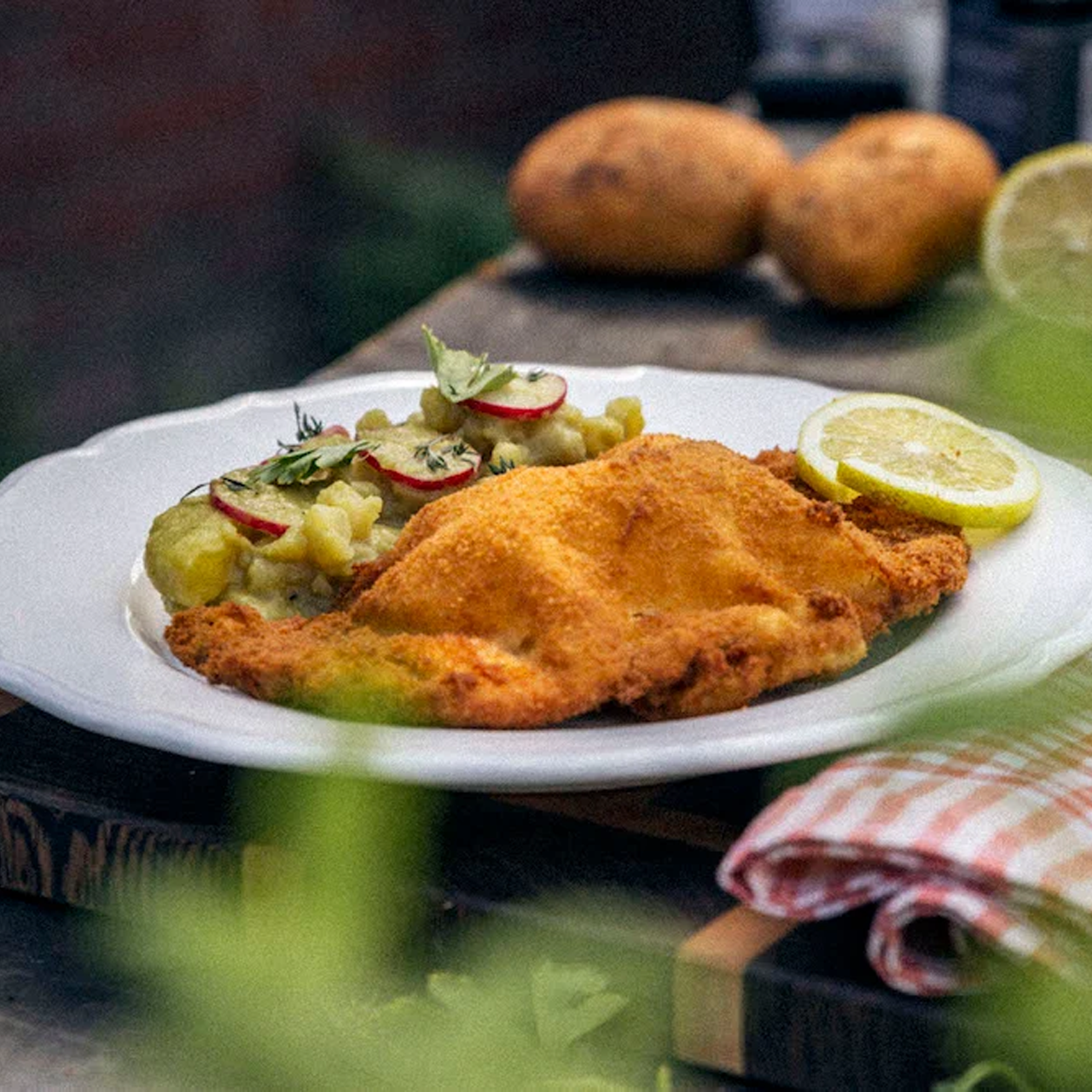 Ein goldbraunes, paniertes Schnitzel mit Zitronenscheibe und Kartoffelsalat, garniert mit Radieschen und Kräutern, auf einem weißen Teller. Im Hintergrund unscharf Kartoffeln und Zitrone.