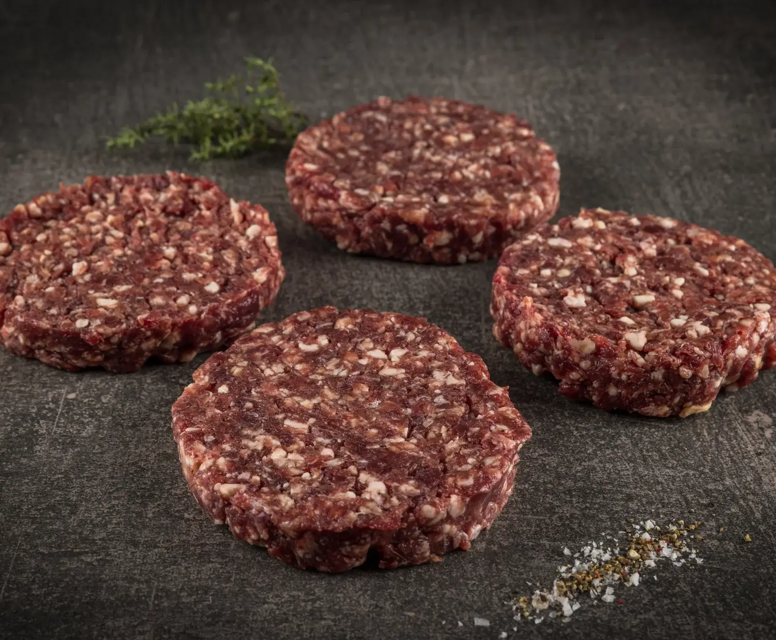 Vier rohe, runde Rinderhackfleisch-Patties liegen auf einer dunklen Steinplatte. Im Hintergrund ist ein Zweig Thymian, im Vordergrund eine Prise Salz und Pfeffer zu sehen.