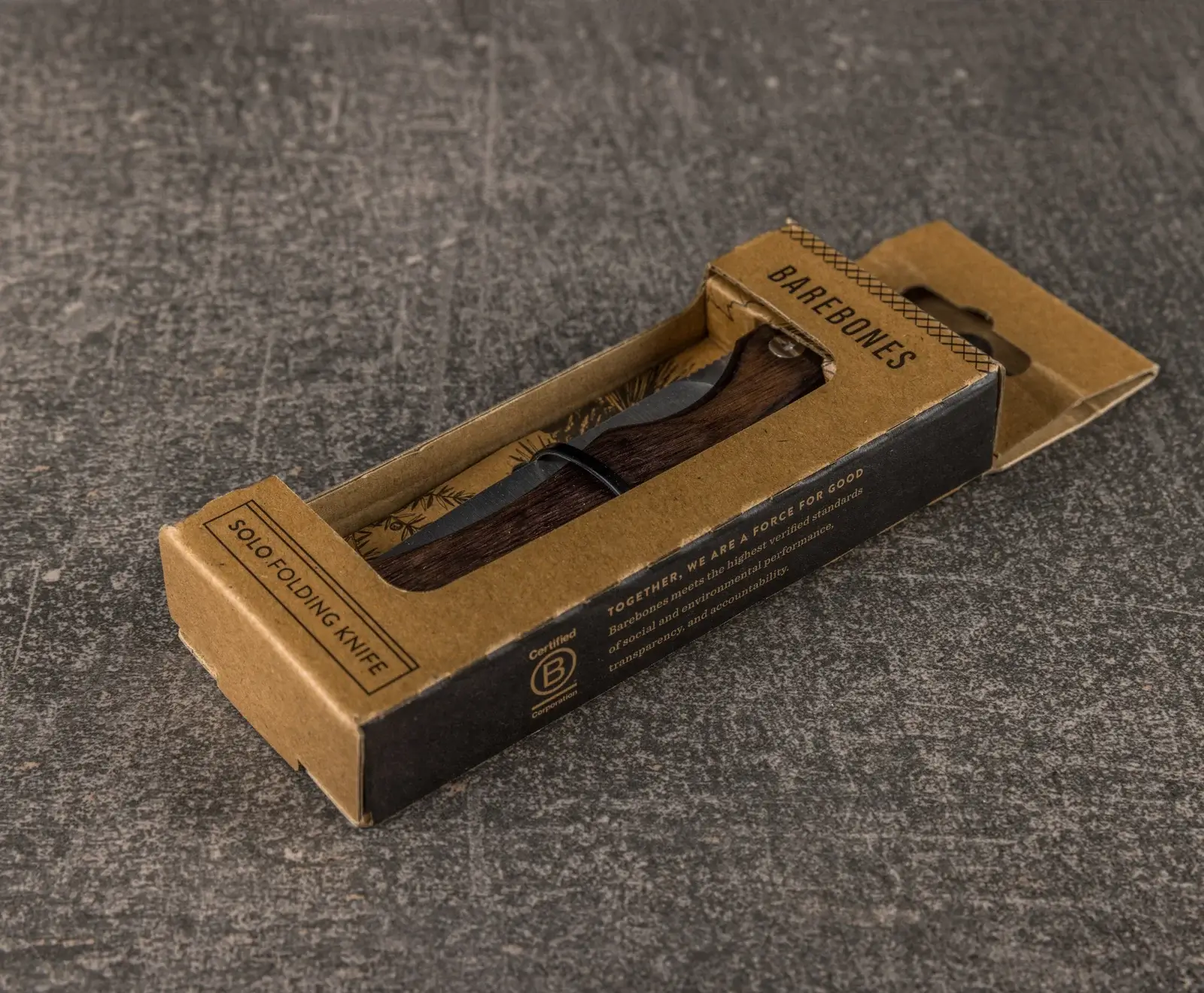 Ein Barebones Solo Klappmesser mit dunklem Holzgriff und dunkler Klinge in seiner braunen Kartonverpackung, die auf einem grauen, strukturierten Untergrund liegt.