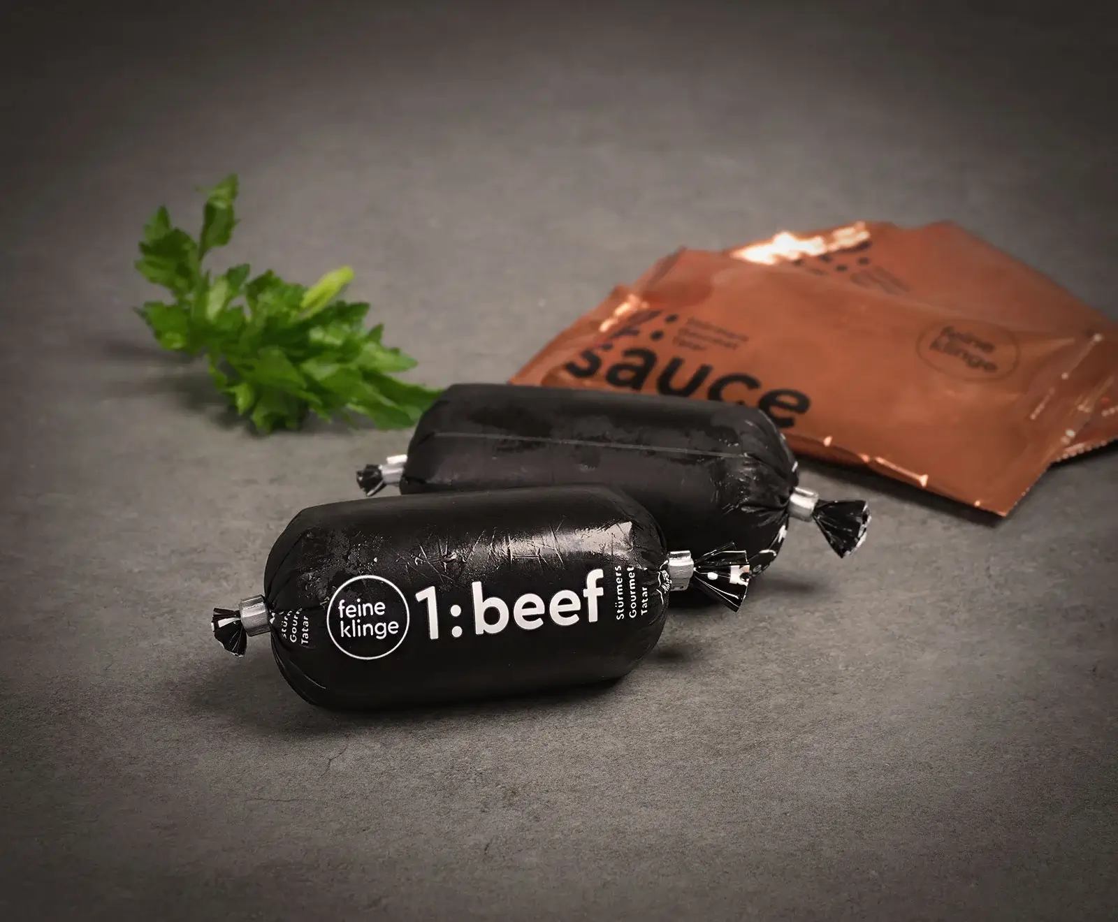Zwei schwarze "feine klinge 1:beef Stürmers Gourmet Tatar" Rollen und zwei kupferfarbene "sauce" Beutel liegen mit einem Petersilienzweig auf dunklem Untergrund.