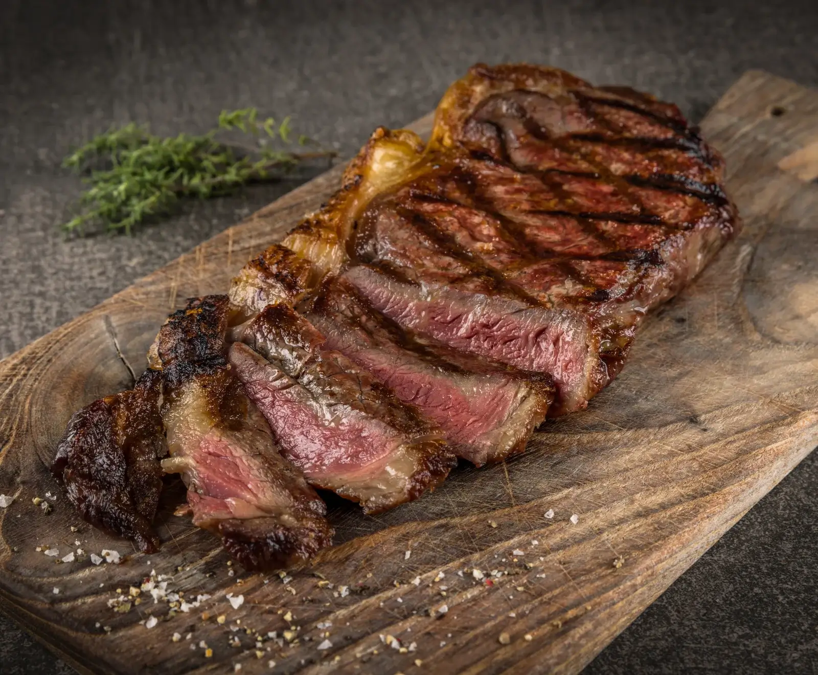 Ein perfekt gegrilltes, teilweise geschnittenes Rib-Eye-Steak auf einem Holzbrett, bestreut mit Salz und Pfeffer, mit Thymian im Hintergrund.
