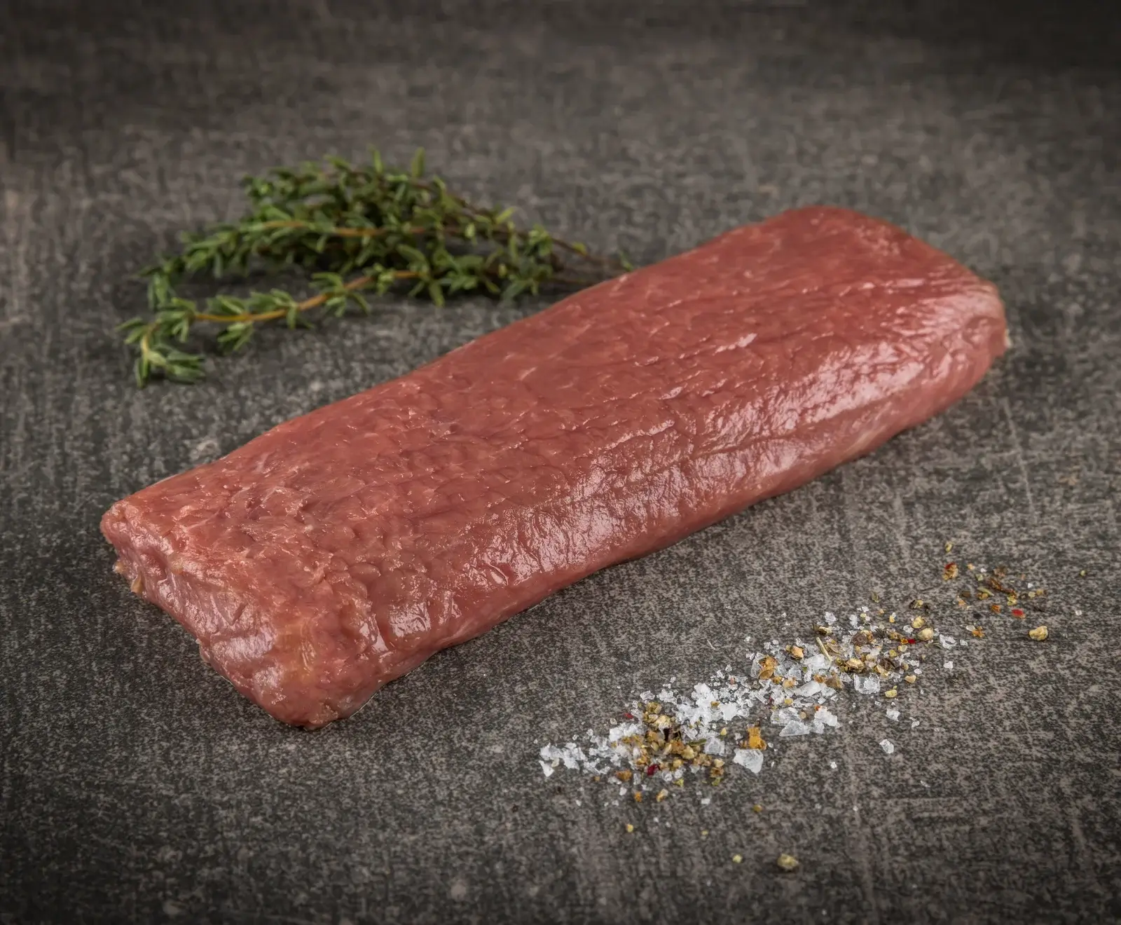 Ein rohes, längliches Stück rotes Fleisch liegt auf einer dunklen Oberfläche, begleitet von einem Thymianzweig und einer Prise grobem Salz und Pfeffer.