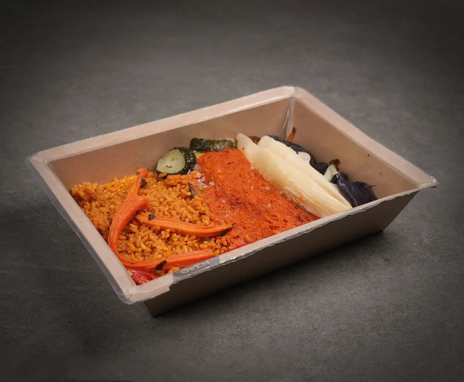 Eine rechteckige, braune Take-away-Schale mit einer Mahlzeit, bestehend aus orangefarbenem Reis mit Karotten, einer roten Sauce, weißen Gemüsestücken, Auberginen und Zucchini, auf dunklem Hintergrund.