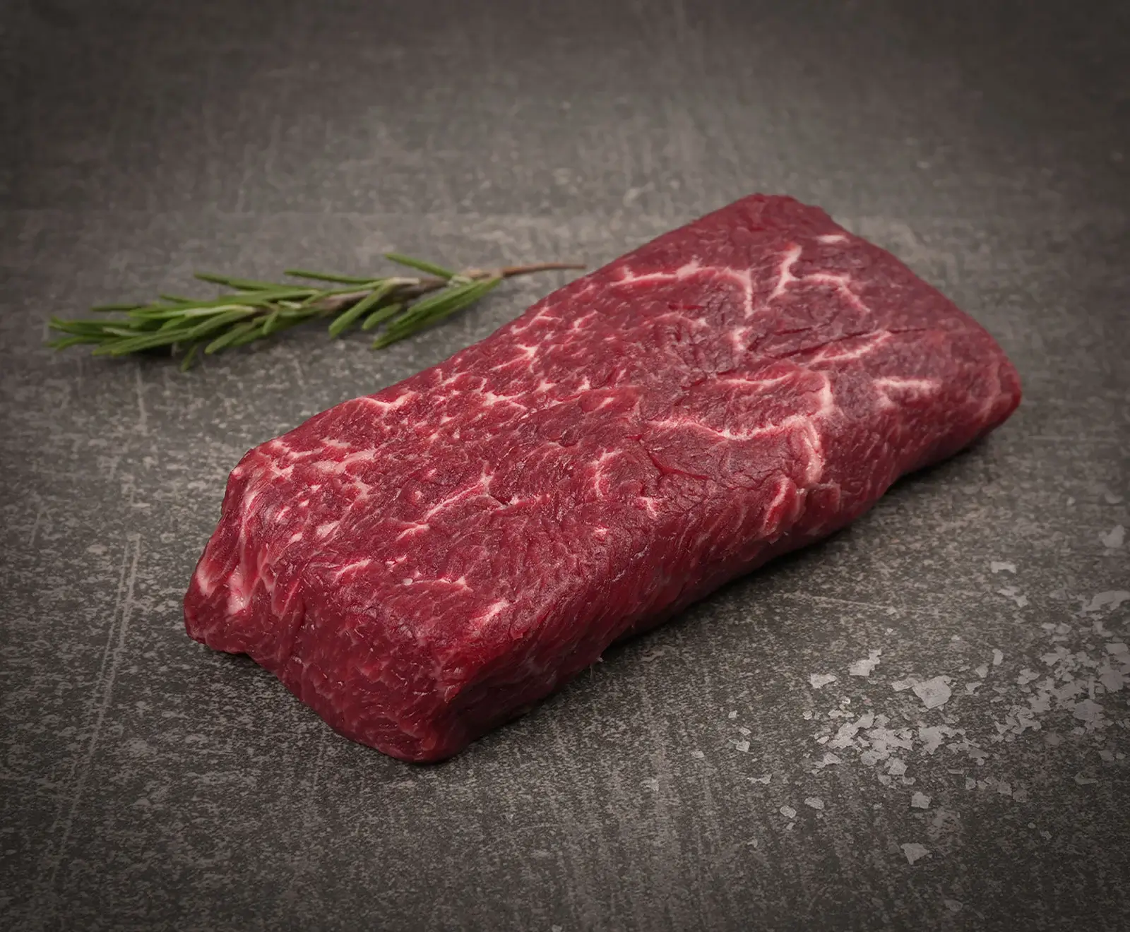 Ein rohes, rechteckiges Stück Rindfleisch mit deutlicher Marmorierung liegt auf einer dunklen Schieferplatte. Links oben befindet sich ein Rosmarinzweig, rechts unten grobe Salzkristalle.
