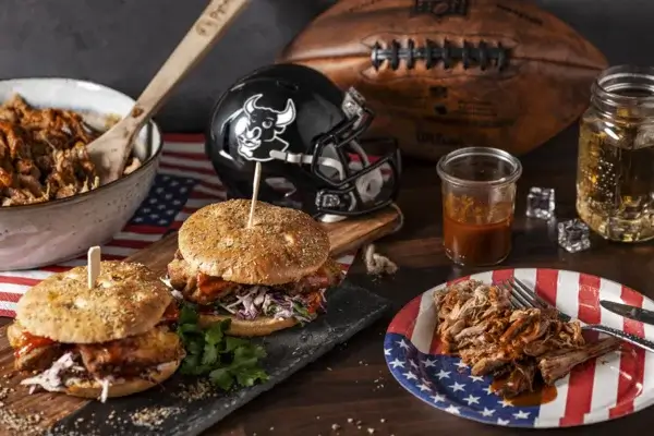 Zwei Pulled Pork Burger mit Krautsalat auf einem Brett, dazu eine Schale und ein Teller mit Pulled Pork. Im Hintergrund sind ein American Football, ein Helm und ein Getränk auf einer US-Flagge zu sehen.
