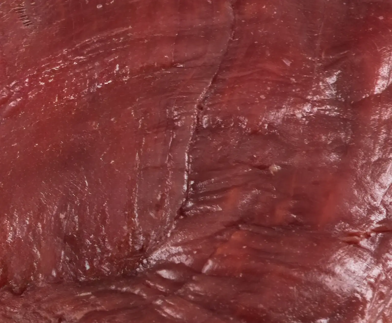 Nahaufnahme von rotem Rohfleisch mit sichtbaren Muskelfasern und einer feuchten, glänzenden Oberfläche. Eine deutliche Falte verläuft vertikal durch die Mitte.