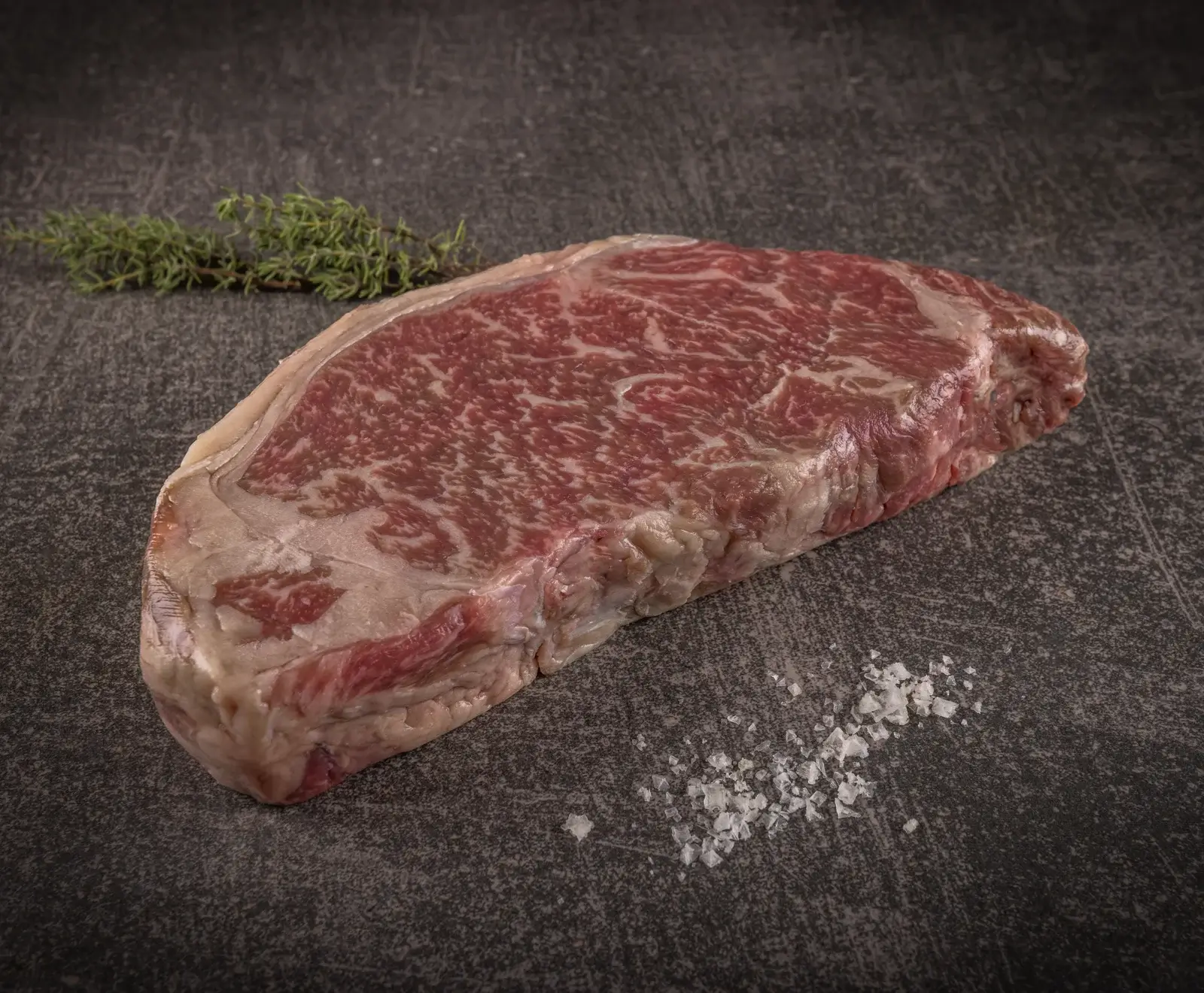 Ein Stück rohes Rindfleisch, marmoriert mit Fett, auf einer dunklen Oberfläche mit Salz daneben.