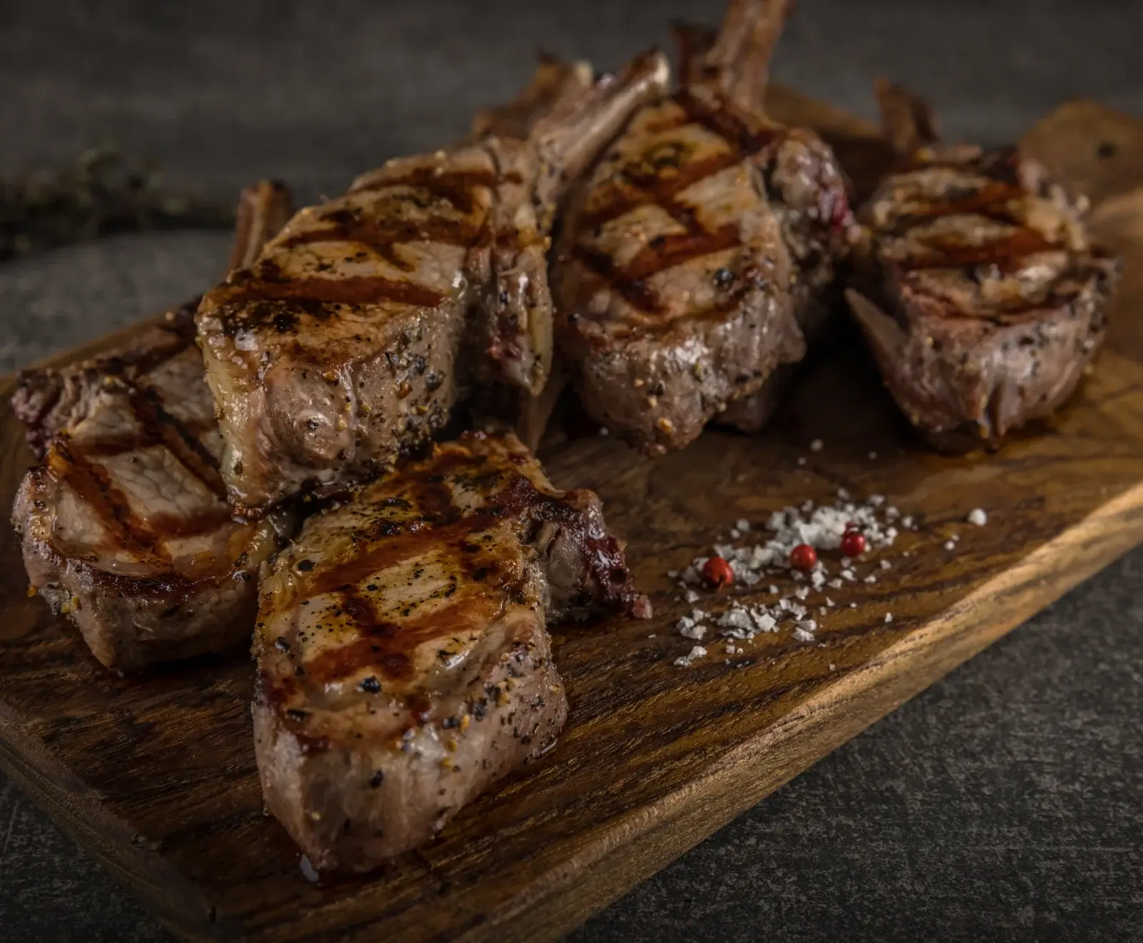 Mehrere gegrillte Lammkoteletts mit deutlichen Grillstreifen und Pfefferwürzung liegen auf einem rustikalen Holzbrett. Daneben sind grobes Salz und rote Pfefferkörner verstreut.