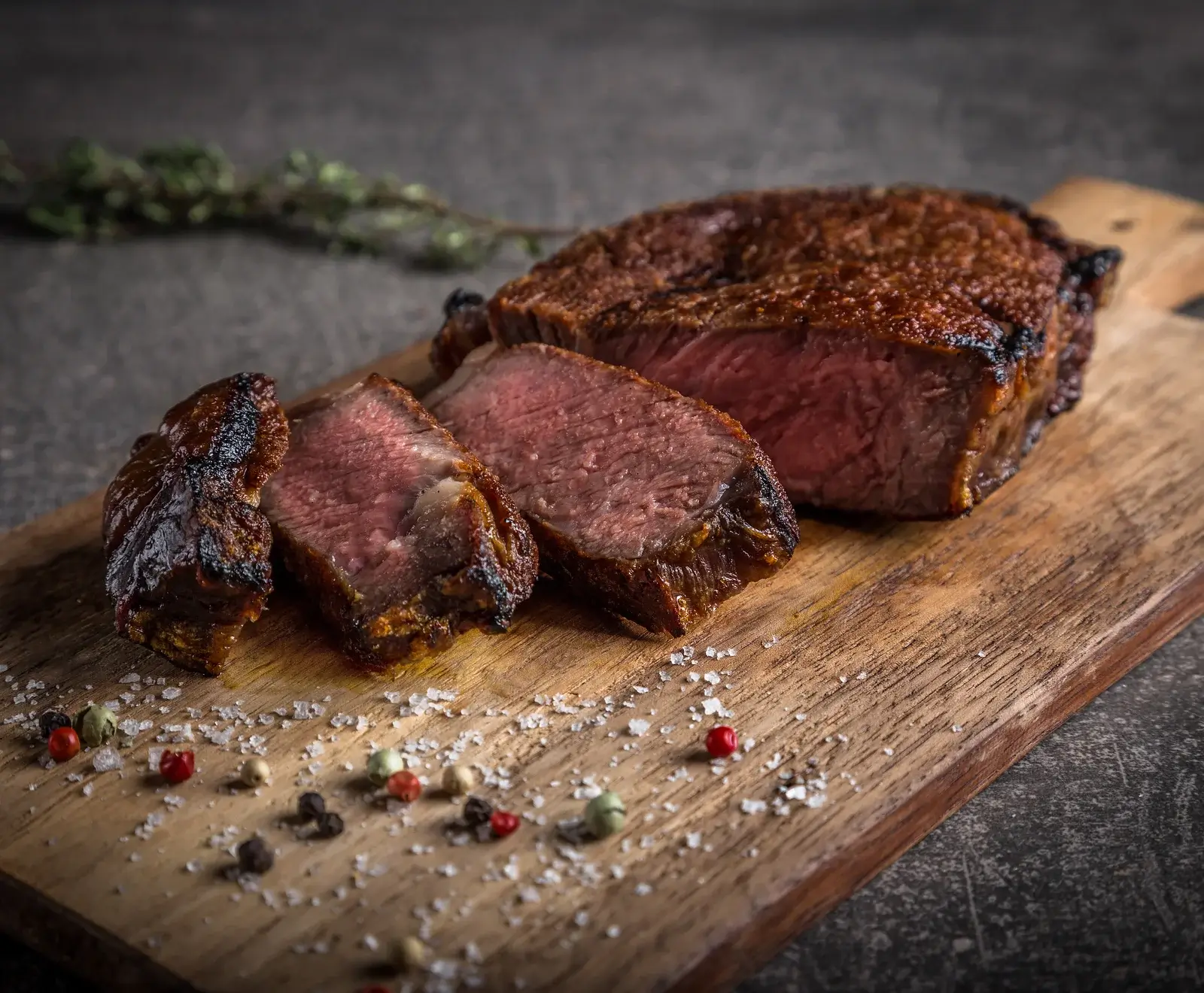 Ein perfekt gegrilltes, teilweise geschnittenes Steak auf einem Holzbrett, umgeben von grobem Salz und bunten Pfefferkörnern. Der Kern ist rosa.