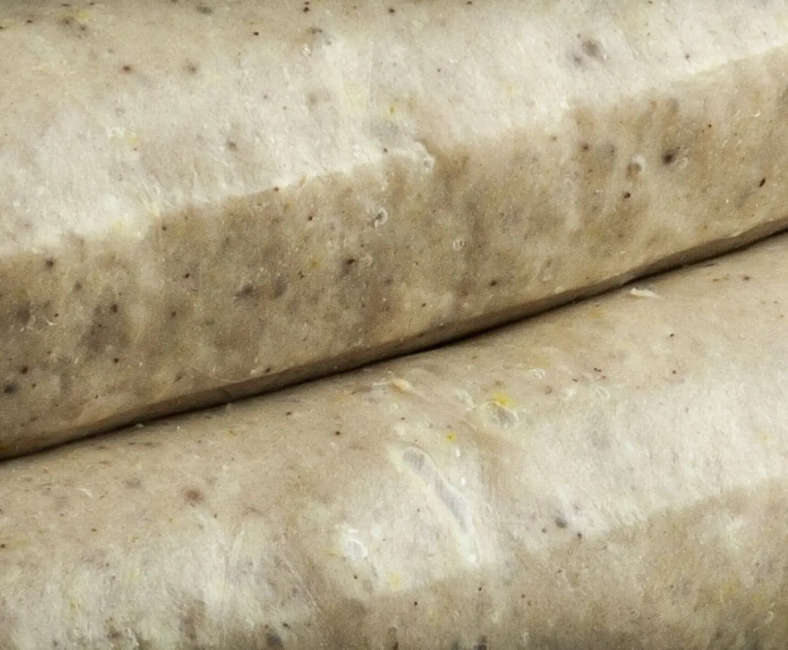 Nahaufnahme von zwei hellen, rohen Würsten, die diagonal übereinander liegen. Die Würste sind zylindrisch, beige-grauweiß und zeigen dunkle Sprenkel von Gewürzen auf ihrer Oberfläche.