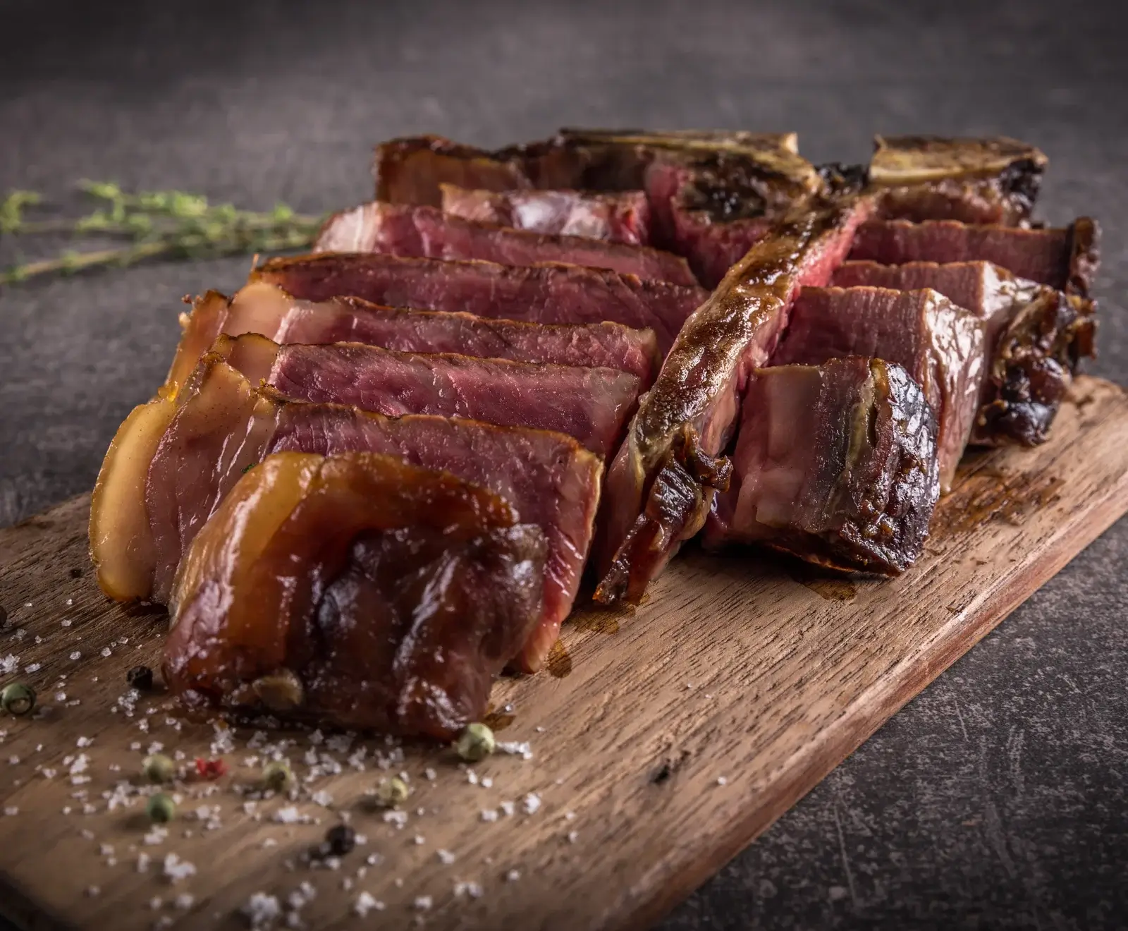 Ein aufgeschnittenes, medium-rare gegartes Steak auf einem Holzschneidebrett, bestreut mit grobem Salz und bunten Pfefferkörnern. Im Hintergrund ein Kräuterzweig.