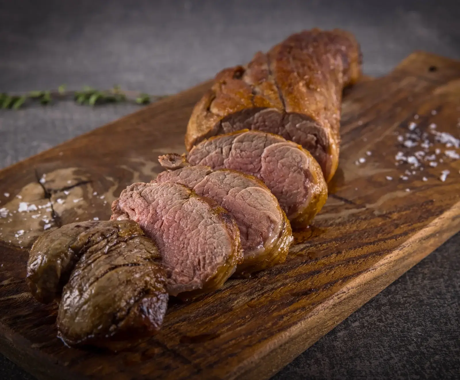 Ein aufgeschnittenes, rosa gebratenes Fleischstück, vermutlich Filet, mit goldbrauner Kruste auf einem rustikalen Holzbrett. Grobe Salzkristalle sind daneben verstreut.