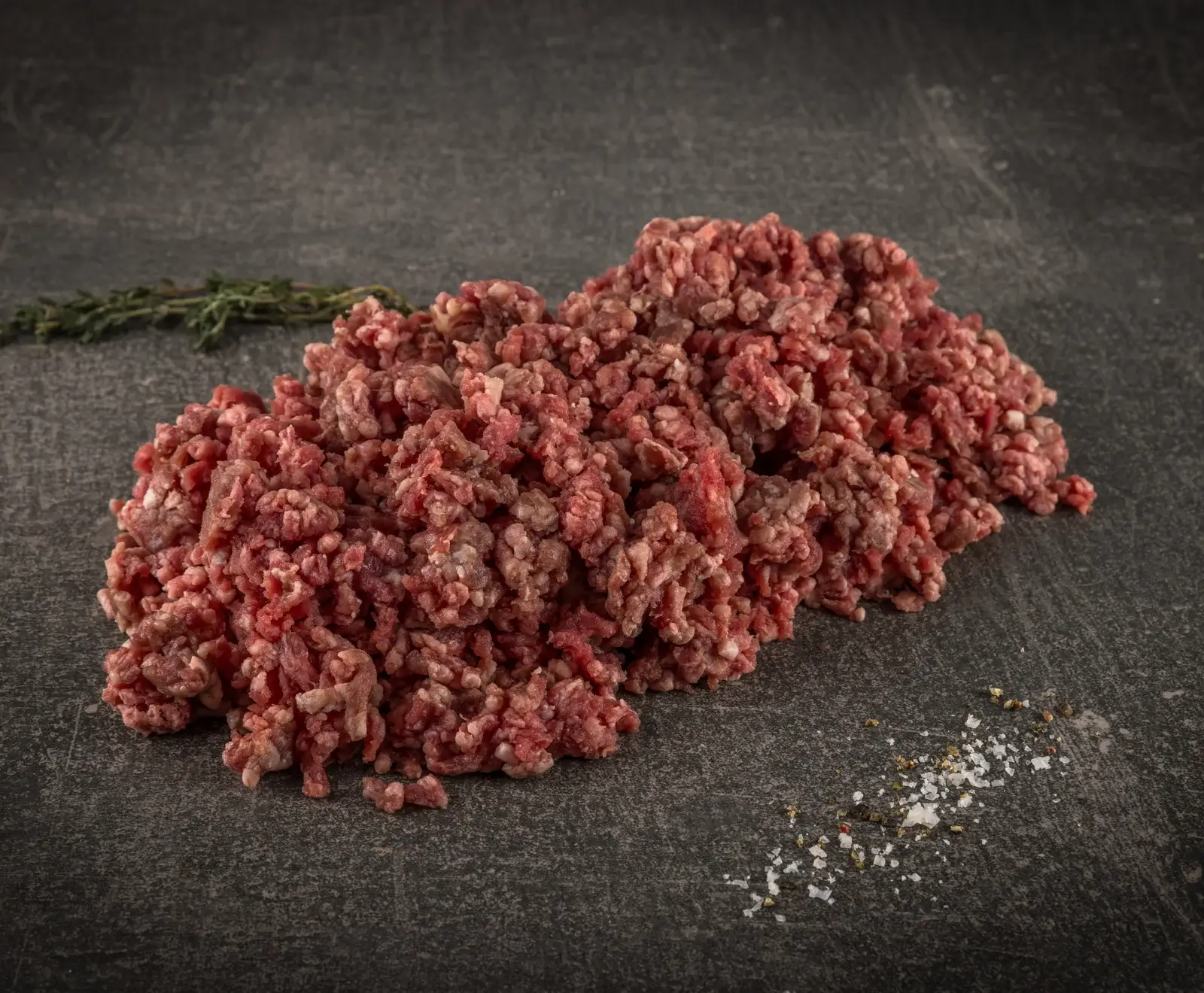 Ein großer Haufen frisches, rohes Hackfleisch liegt auf einer dunklen, strukturierten Oberfläche. Links im Hintergrund ist ein Thymianzweig, rechts im Vordergrund grobes Salz und Pfeffer verstreut.