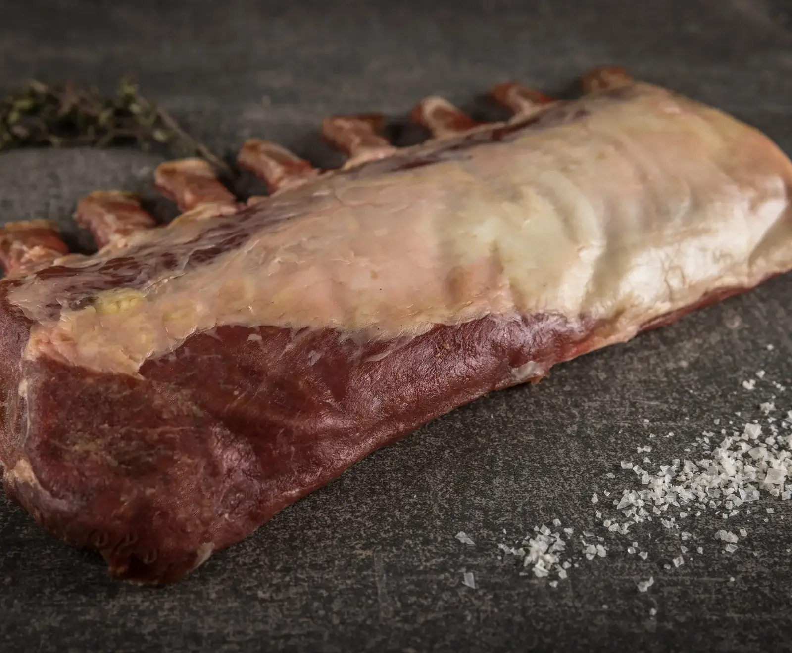 Ein rohes Lammkarree mit sichtbaren Knochen und einer Fettschicht liegt auf einer dunklen, strukturierten Oberfläche. Im Hintergrund sind Thymianzweige, im Vordergrund grobes Meersalz verstreut.