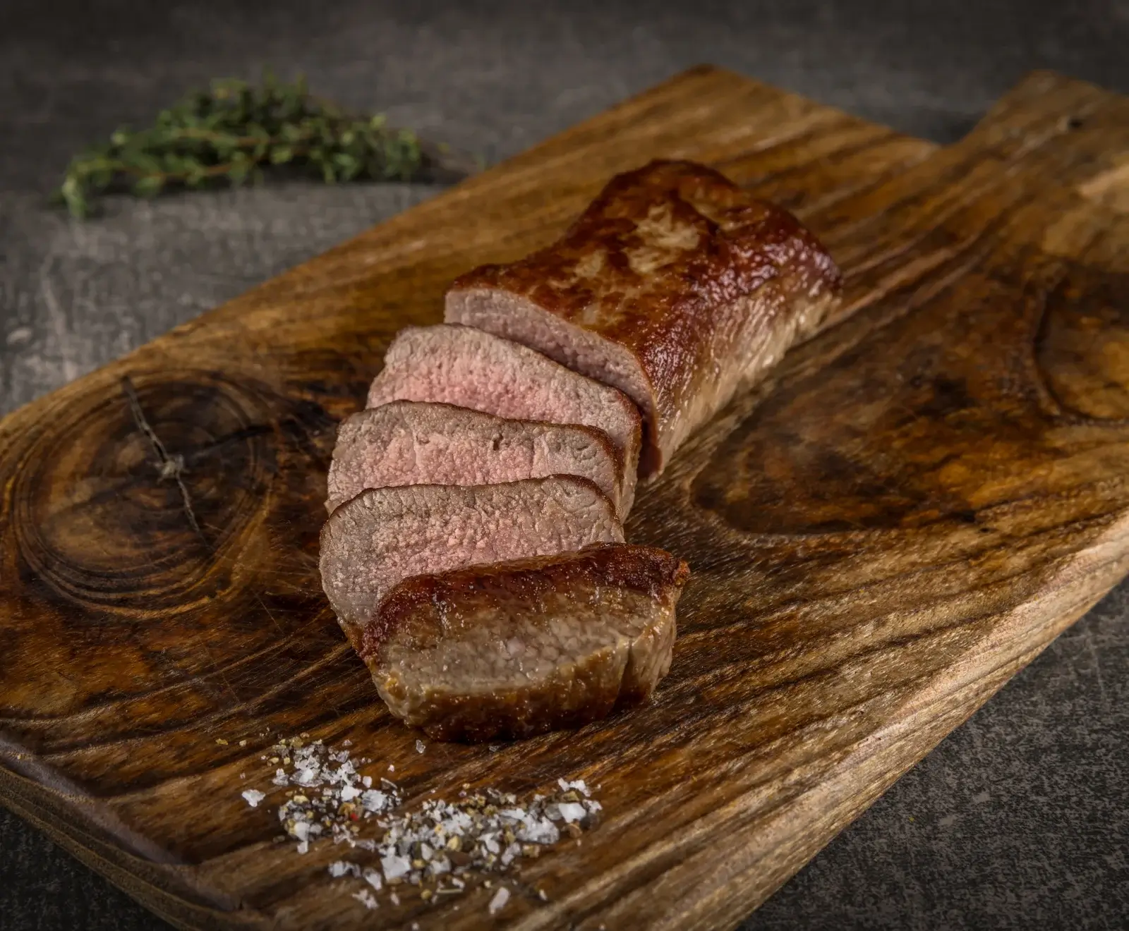 Ein aufgeschnittenes, medium gebratenes Stück Rinderfilet oder Lende auf einem rustikalen Holzbrett, garniert mit grobem Salz, Pfeffer und einem Thymianzweig.