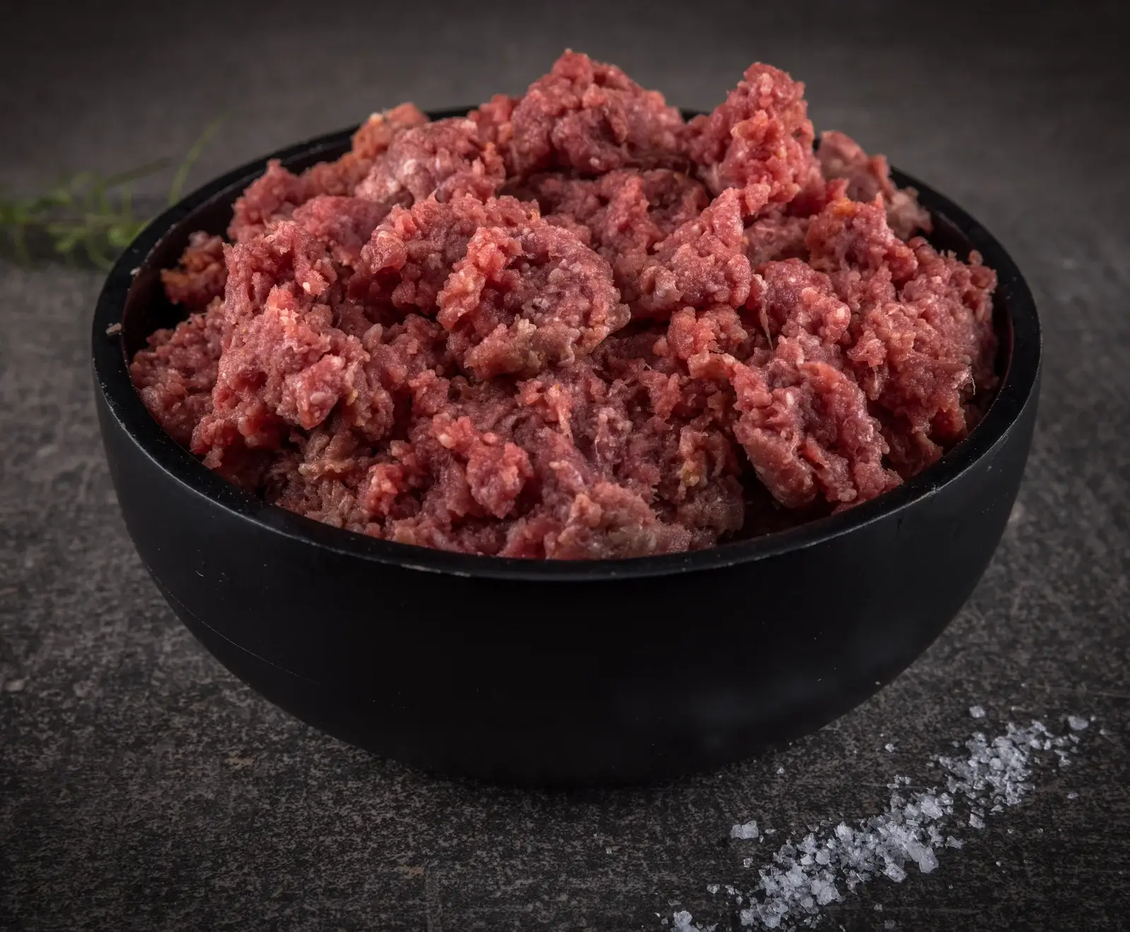 Eine schwarze Schale, gefüllt mit frischem, rotem Hackfleisch, steht auf einer dunklen, strukturierten Oberfläche. Rechts daneben liegt grobes Salz, links im Hintergrund ist ein Rosmarinzweig zu sehen.