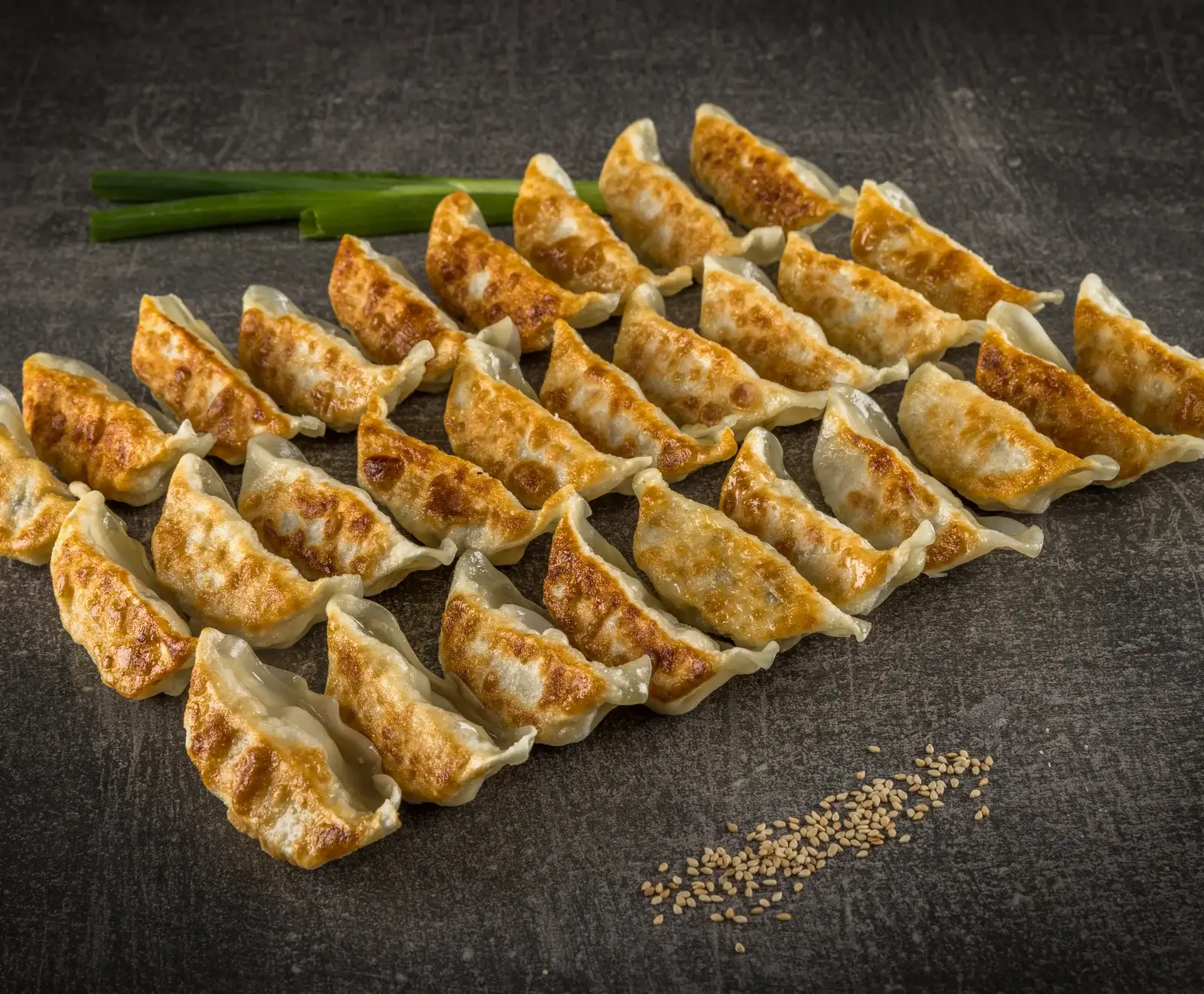 Eine große Menge goldbraun gebratener Gyoza-Teigtaschen, ordentlich in Reihen auf einer dunklen Oberfläche angeordnet, mit zwei Lauchzwiebeln und Sesamsamen im Hintergrund.