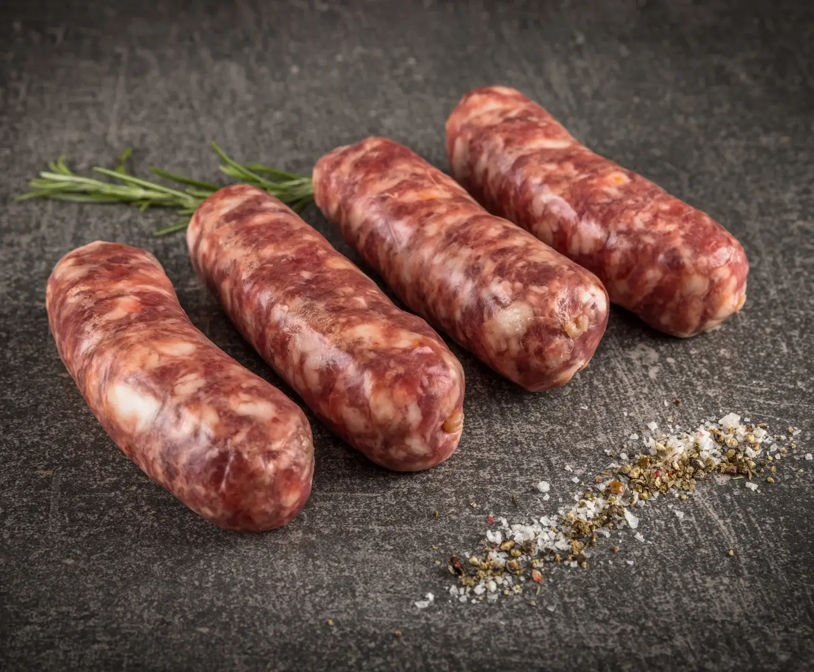 Vier rohe, grobe Bratwürste liegen auf einer dunklen Oberfläche, daneben Rosmarinzweige und eine Prise Salz und Pfeffer.