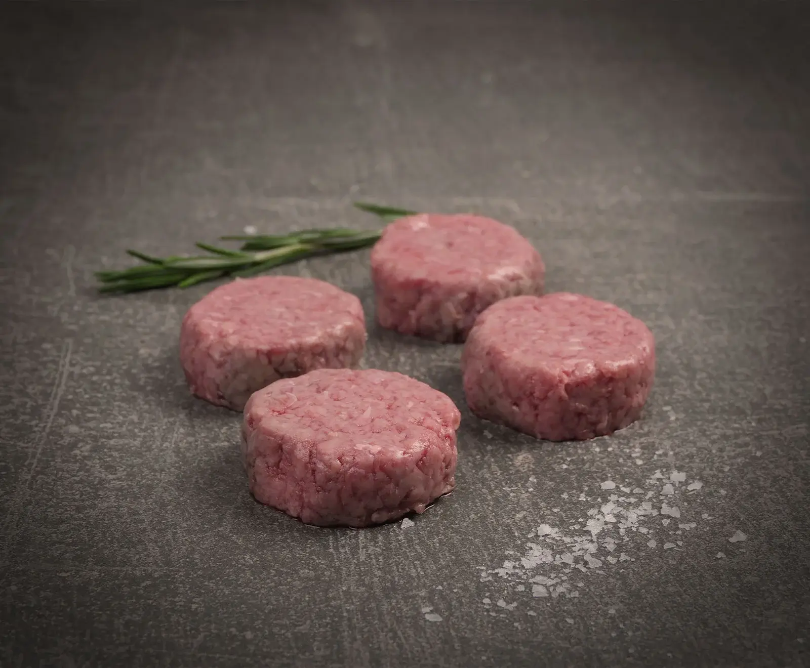 Vier rohe, runde Fleischpatties liegen auf einer dunklen Oberfläche. Im Hintergrund ist ein Rosmarinzweig, im Vordergrund grobes Salz verstreut.