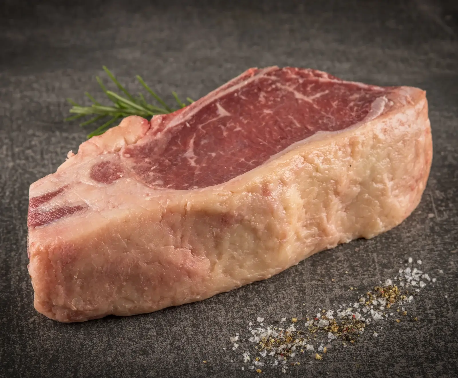 Ein rohes Rindersteak mit deutlichem Fettrand und feiner Marmorierung liegt auf einem dunklen Untergrund. Daneben sind ein Rosmarinzweig sowie verstreute Salz- und Pfefferkörner zu sehen.
