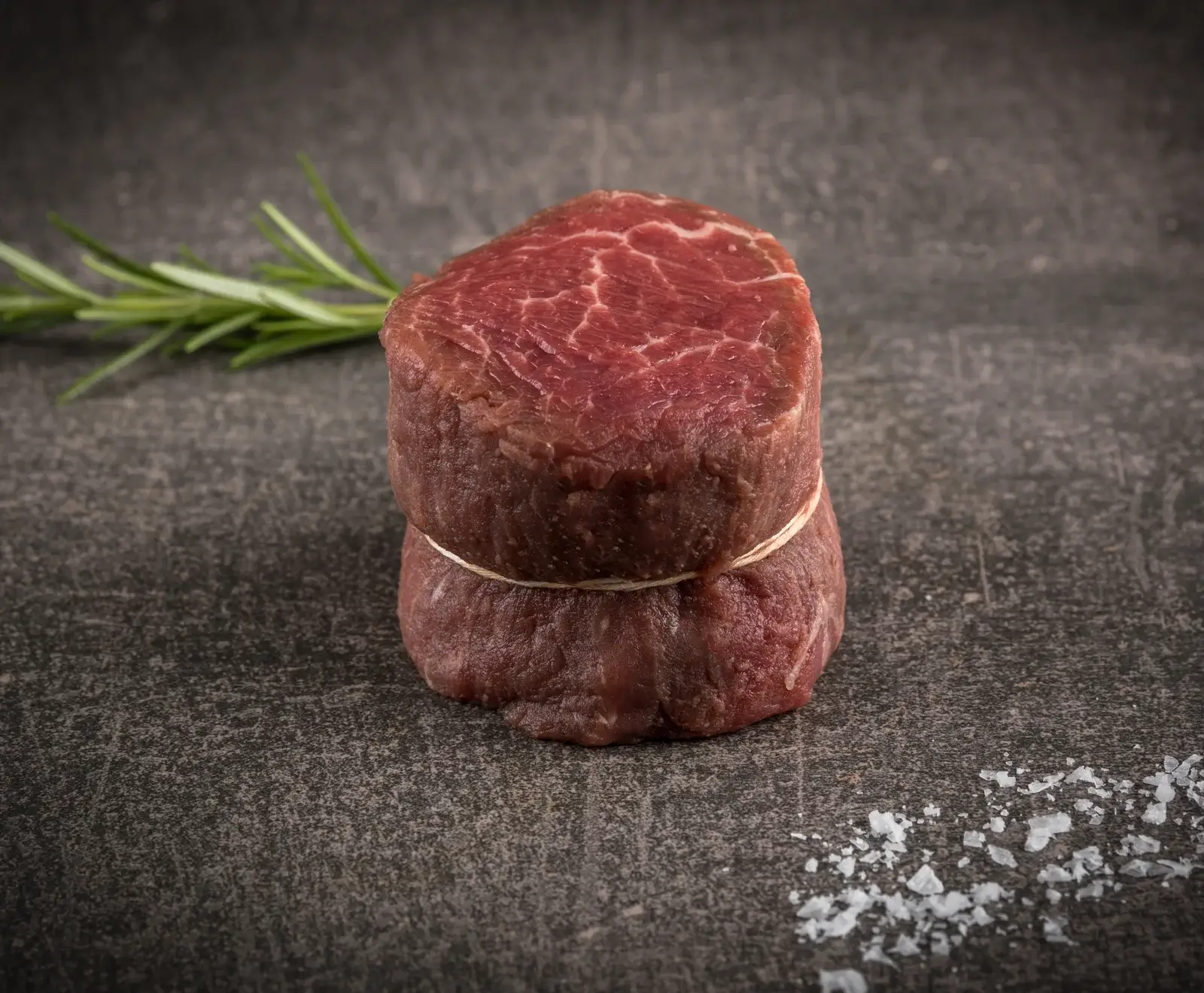 Ein rohes, mit Küchengarn gebundenes Rinderfiletmedaillon liegt auf einer dunklen, strukturierten Oberfläche. Links daneben ein Rosmarinzweig, rechts unten grobes Meersalz.