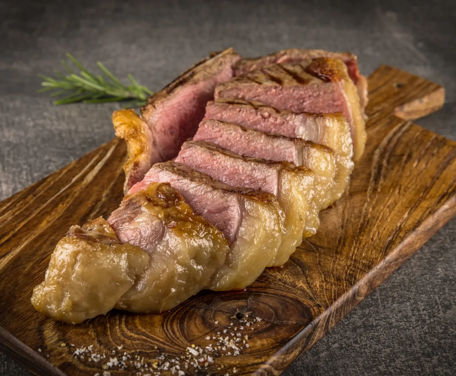 Ein perfekt gegrilltes, in Scheiben geschnittenes Steak mit rosa Kern und goldbrauner Kruste liegt auf einem Holzschneidebrett. Im Hintergrund ist ein Rosmarinzweig zu sehen, im Vordergrund grobes Salz.