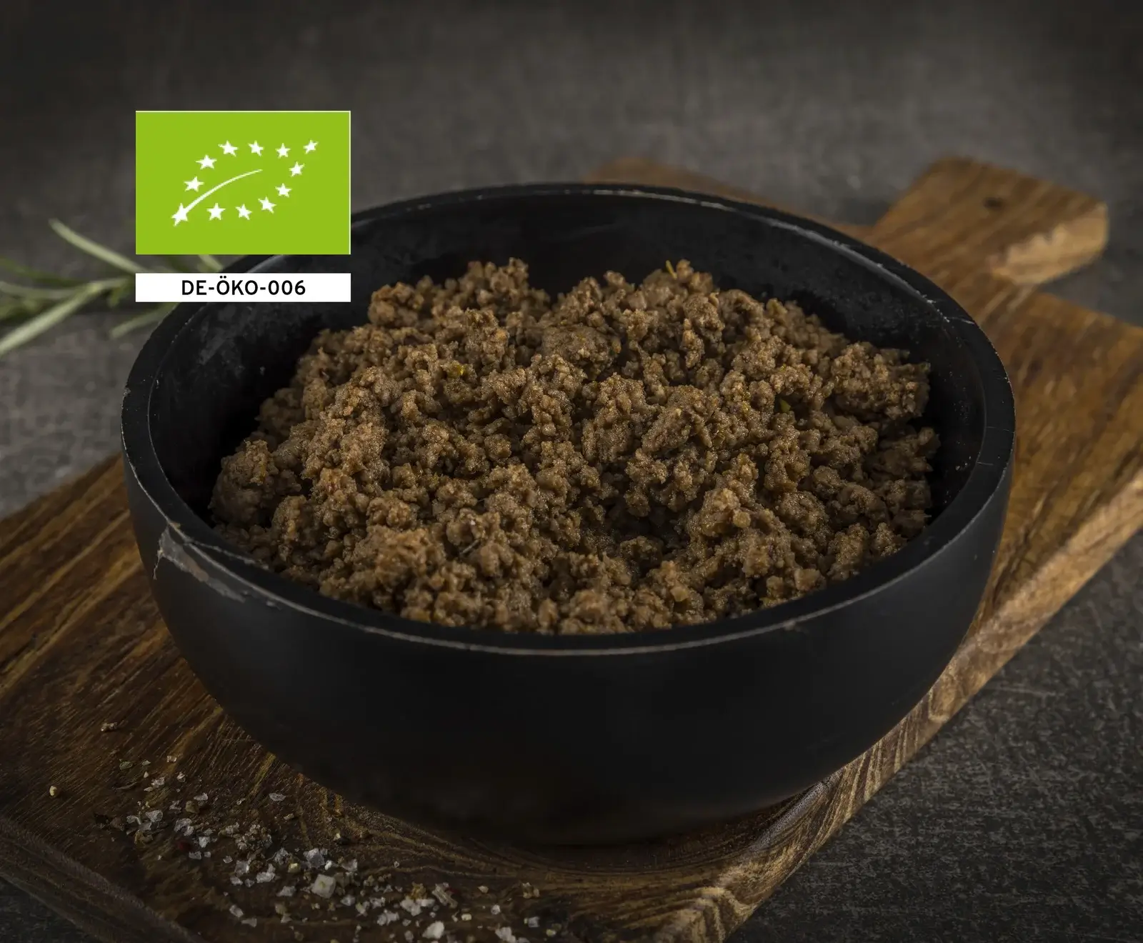 Schwarze Schüssel gefüllt mit braunen, krümeligen Flocken auf einem Holzbrett, daneben ein grünes EU-Bio-Logo.