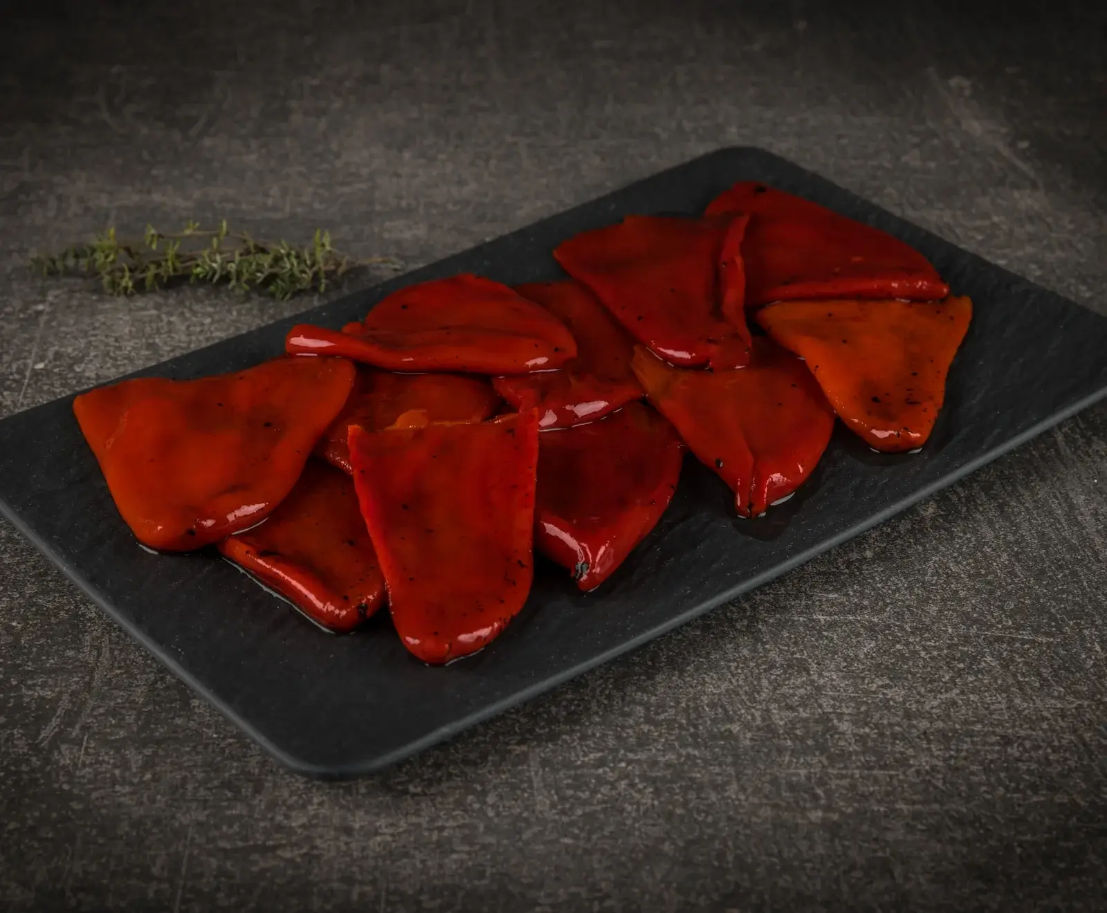 Glänzende, geröstete rote Paprikastreifen liegen auf einer schwarzen rechteckigen Platte. Im Hintergrund ist ein kleiner Zweig frischer Thymian zu sehen.