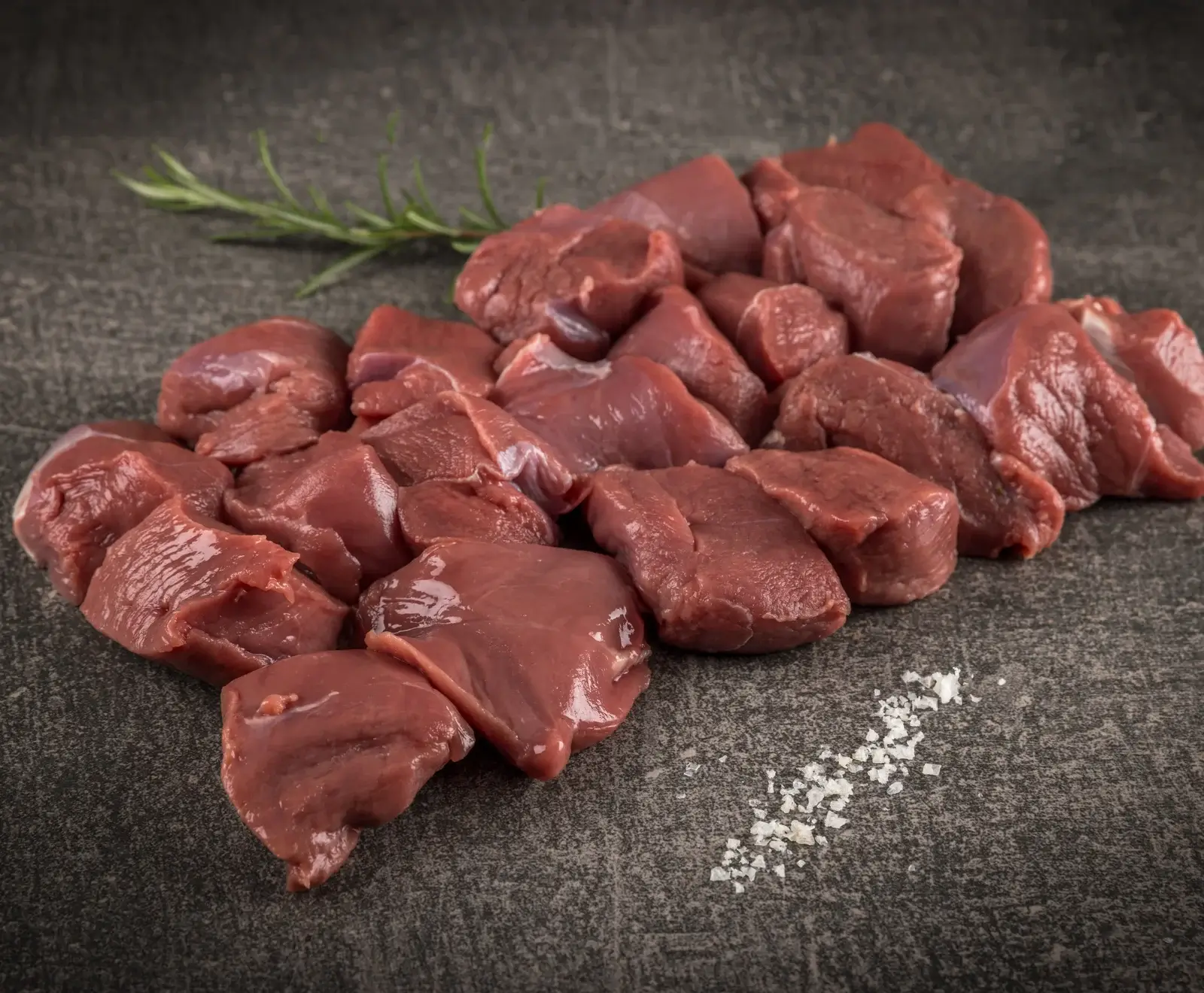 Ein Haufen roher, dunkelroter Fleischwürfel, möglicherweise Wildfleisch, liegt auf einer dunklen, strukturierten Oberfläche. Ein Rosmarinzweig und grobe Salzkristalle sind daneben platziert.