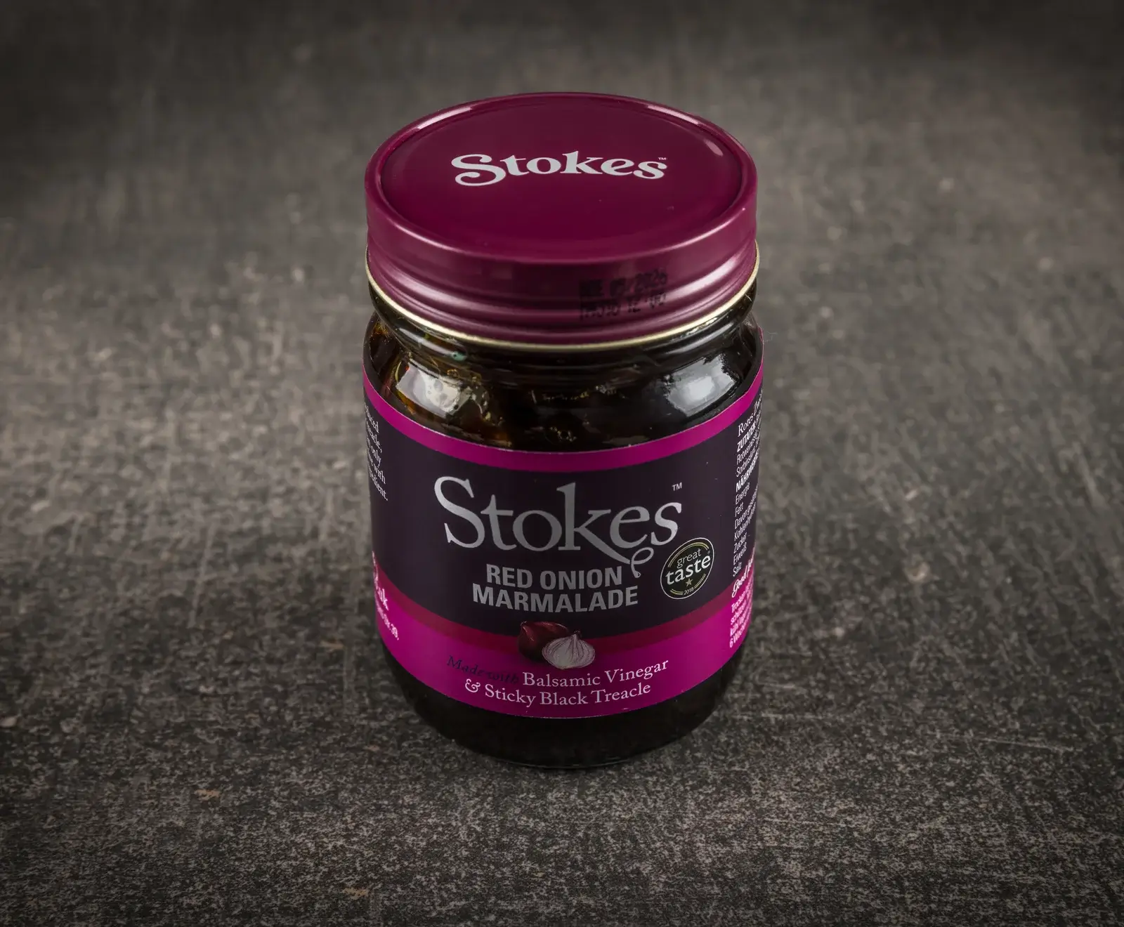 Ein Glas Stokes Red Onion Marmalade mit lila Etikett und Deckel, das Balsamico-Essig und schwarzen Sirup enthält, auf dunklem, strukturiertem Untergrund.
