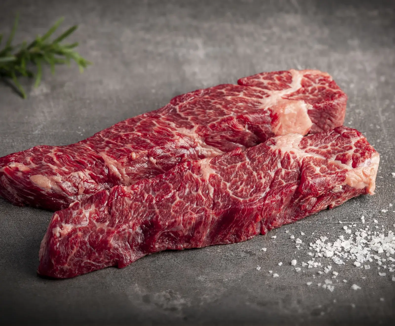 Drei rohe Wagyu-Rindfleisch-Steaks mit Meersalz auf einer dunklen Schieferplatte mit Rosmarin garniert.