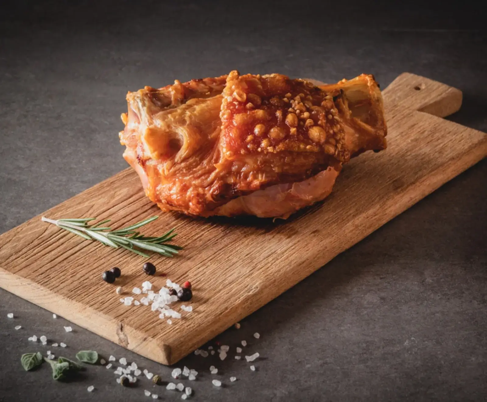 Eine goldbraun gebratene Schweinshaxe mit knuspriger Haut auf einem rustikalen Holzschneidebrett, umgeben von Rosmarin, Pfefferkörnern und Meersalz.