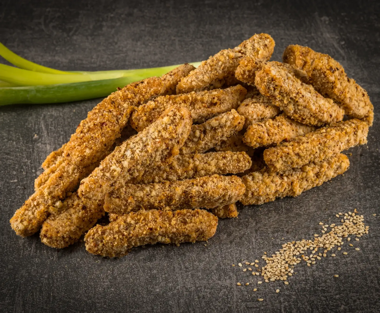 Goldbraune, knusprig panierte Sticks, daneben eine grüne Frühlingszwiebel und verstreute Sesamsamen auf dunklem Stein.