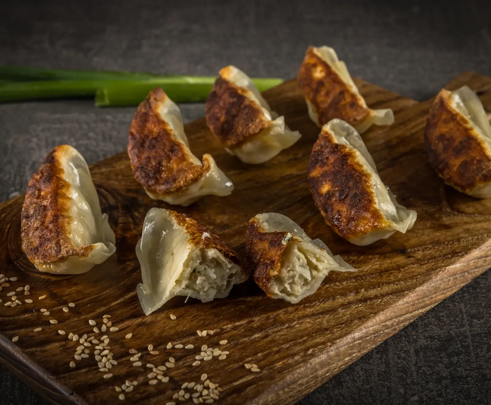 Mehrere goldbraun gebratene Gyoza auf einem Holzbrett, bestreut mit Sesamsamen. Eine Gyoza ist halbiert und zeigt die helle Füllung. Im Hintergrund ist eine grüne Zwiebel zu sehen.
