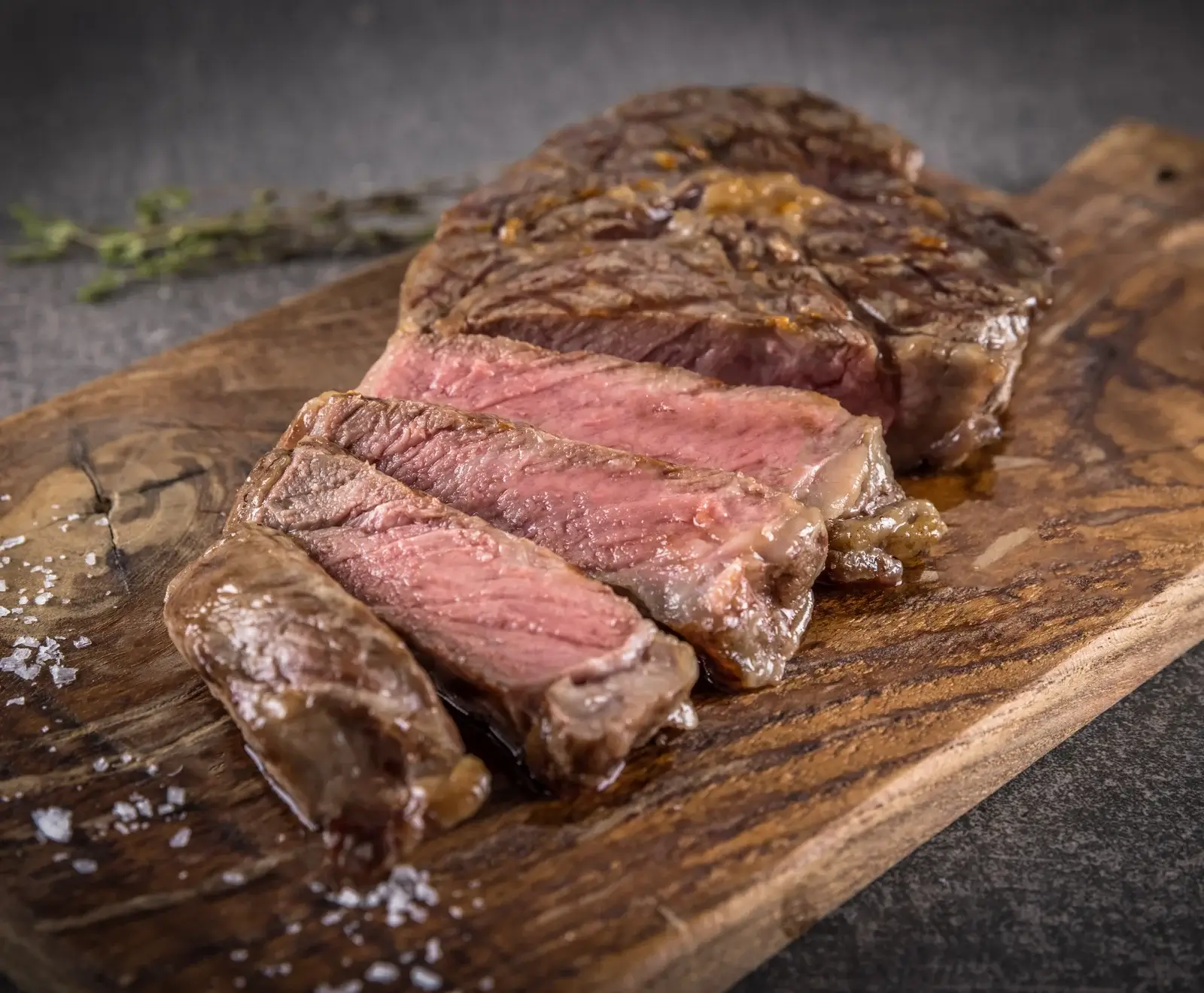 Ein medium-rare gegartes, in Scheiben geschnittenes Steak liegt auf einem rustikalen Holzbrett, umgeben von grobem Salz und einem Kräuterzweig im Hintergrund.