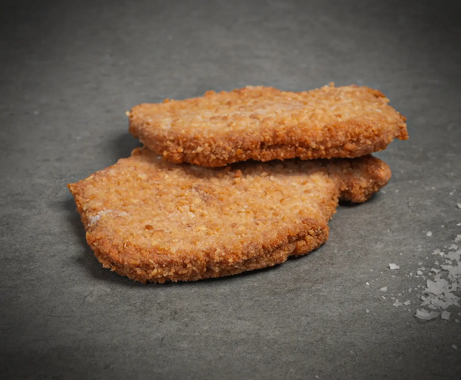 Zwei ungekochte, panierte Schnitzel liegen übereinander auf einer dunkelgrauen Oberfläche. Rechts sind einige grobe Salzkristalle verstreut. Die Panade ist goldbraun.