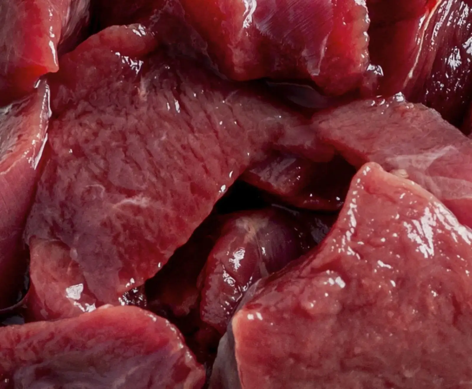 Nahaufnahme von rohem, in Stücke geschnittenem Fleisch. Die tiefroten Fleischstücke sind feucht und glänzen leicht, mit sichtbaren Muskelfasern und einigen helleren Stellen.