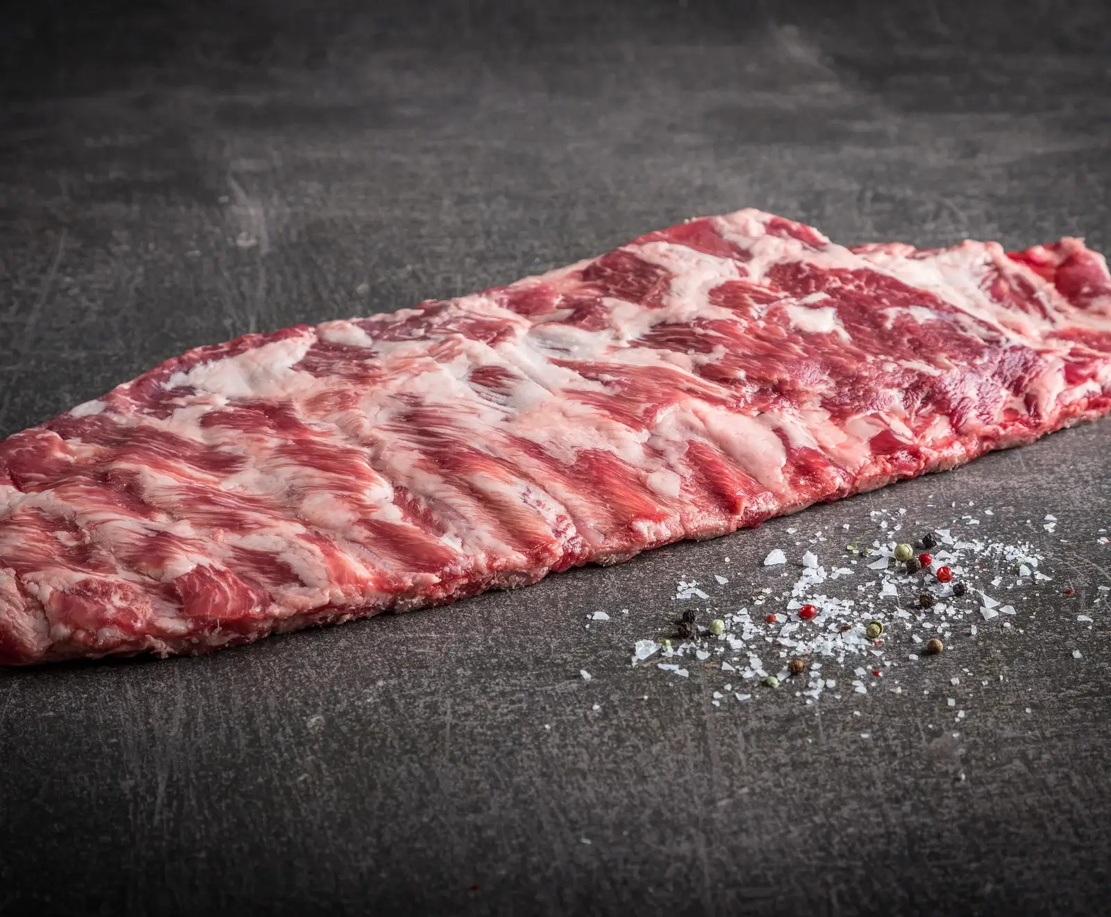Ein großes, rohes Rippchenstück (Spareribs) mit viel Marmorierung liegt auf einer dunklen Oberfläche. Daneben sind grobes Salz und bunte Pfefferkörner verstreut.