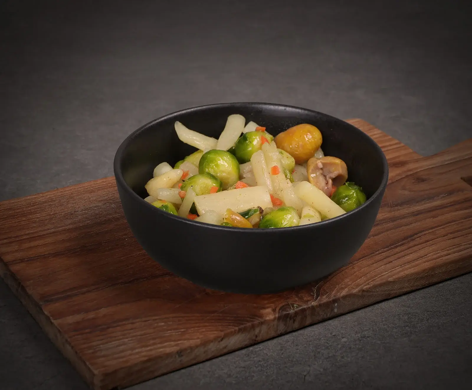 Eine schwarze Schale gefüllt mit einem Gericht aus Rosenkohl, weißem Spargel und Karottenstücken, angerichtet auf einem rustikalen Holzbrett vor dunklem Hintergrund.