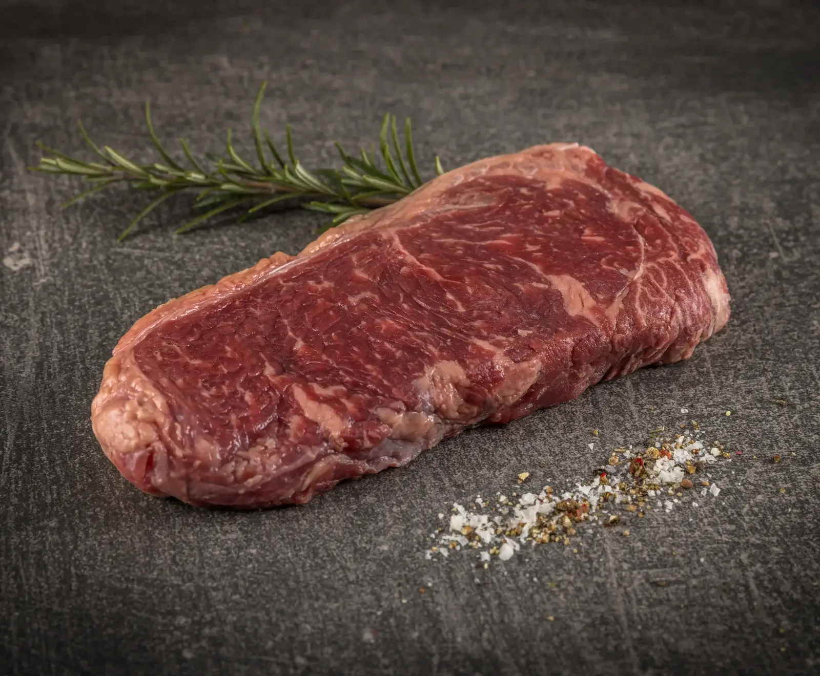 Ein rohes, marmoriertes Rindersteak liegt auf einem dunklen Untergrund, daneben ein Rosmarinzweig und eine Prise grobes Salz mit Pfeffer.