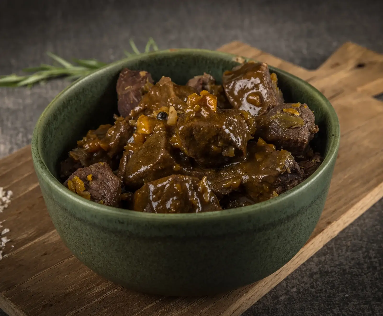 Herzhafter Fleischgulasch in einer dunkelgrünen Keramikschale, serviert auf einem rustikalen Holzbrett. Die dunkle Soße enthält Fleischstücke und Gemüse. Im Hintergrund sind Rosmarin und grobes Salz zu sehen.